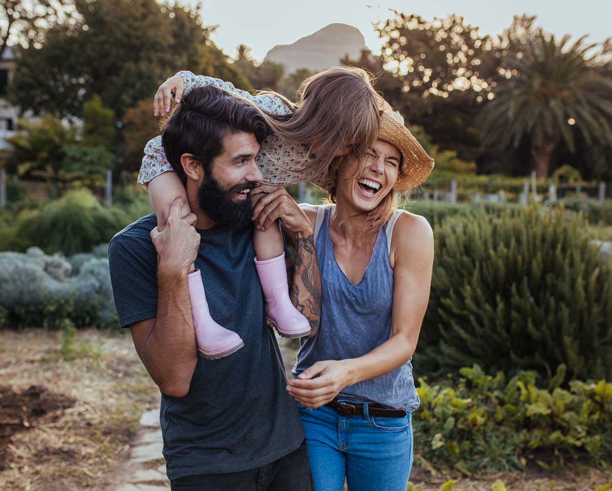 girl sitting on her dads shoulders while mom and dad are laughing, outside on a farm